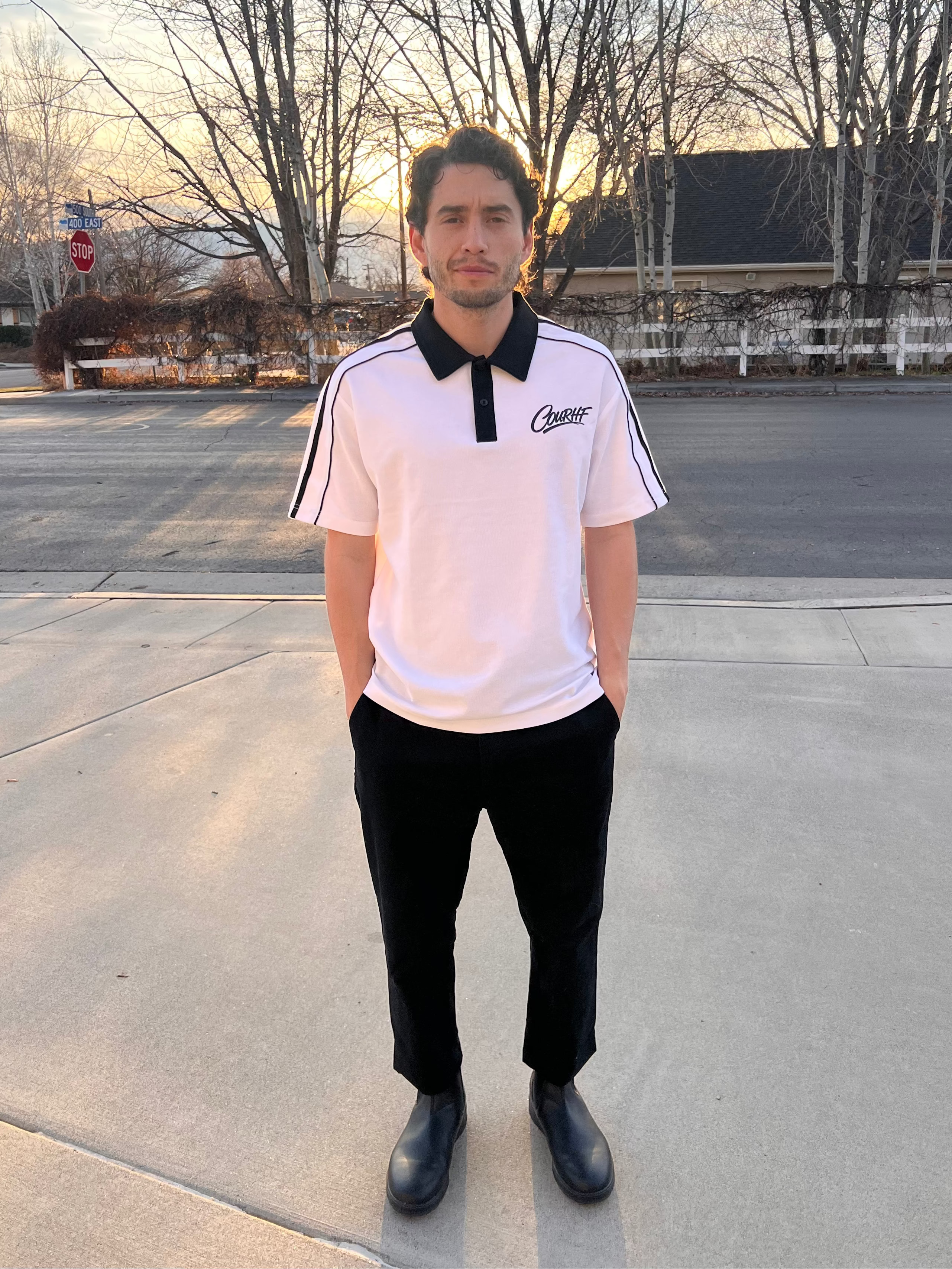 Person wearing a white polo shirt with black accents standing on a sidewalk.